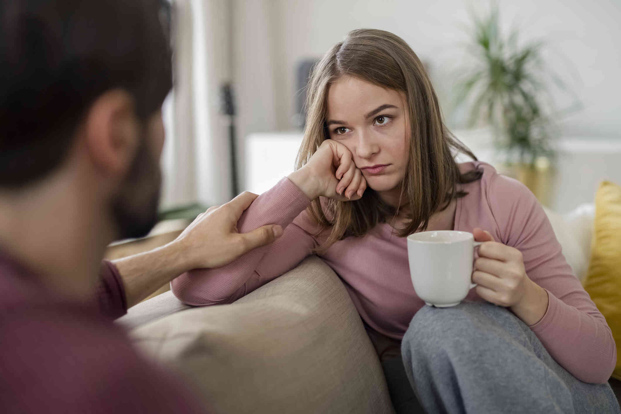 A woman with a coffee mug sits on the couch while rsting her head on her hand with a sad expression as her partner reaches out to touch her arm.